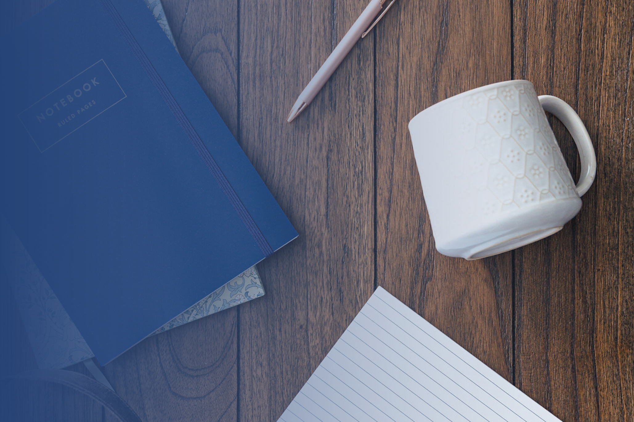 A blue notebook, a pink pen, a white patterned mug, and a lined sheet of paper are arranged on a wooden surface—perfect for jotting down thoughts during family therapy or couples therapy sessions.