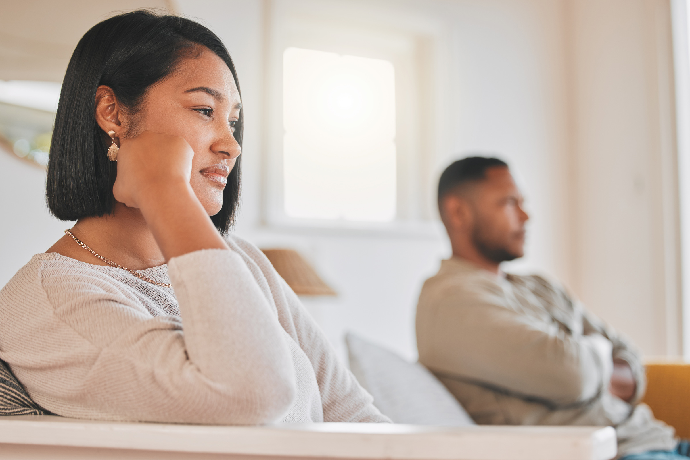 A woman sits in the foreground with her chin resting on her hand, looking thoughtful. A man sits in the background, arms crossed, both appearing distant on a couch—an image reflecting moments before couples therapy in York.
