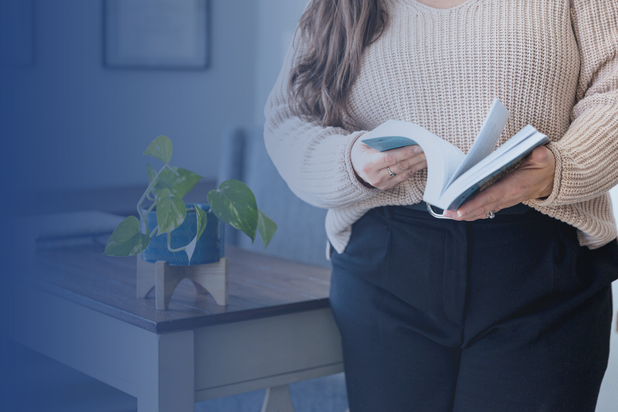 A person in a beige sweater and black pants stands by a table with a potted plant, flipping through a book on family therapy. The softly blurred background adds a calm, focused atmosphere.