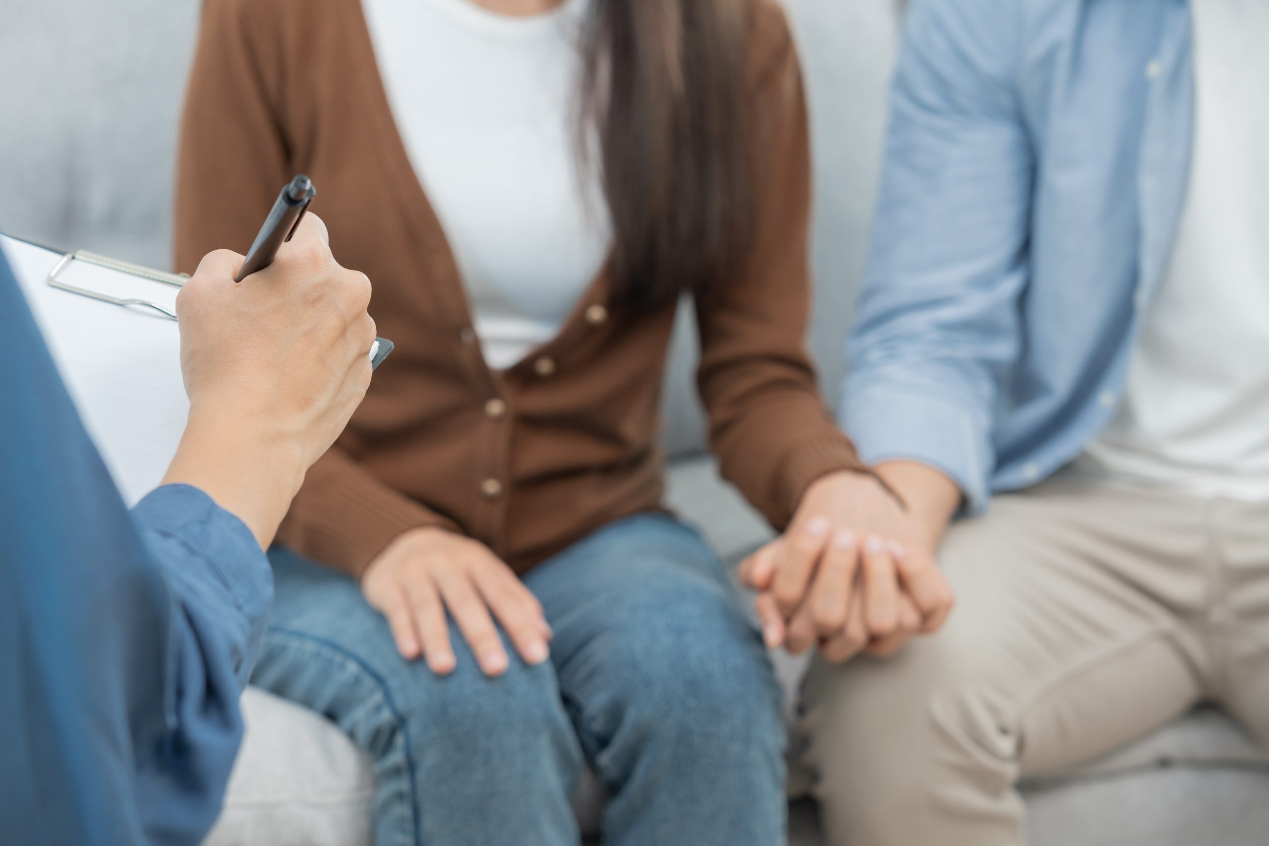 A couple sits closely together, holding hands, while talking to a counselor from Therapeutic Family Connections who is taking notes during a family therapy session. The focus is on their hands and the counselor’s writing hand.