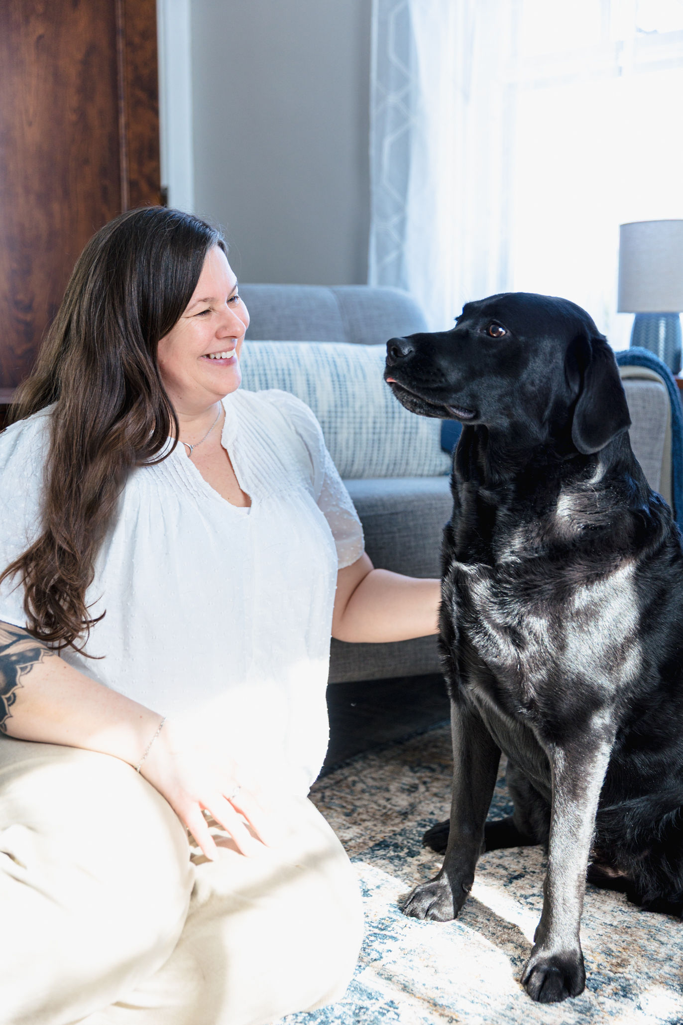 A woman with long brown hair and a white shirt sits on the floor, smiling at a large black dog beside her in a bright, cozy living room—a peaceful space perfect for family therapy or Therapeutic Family Connections.