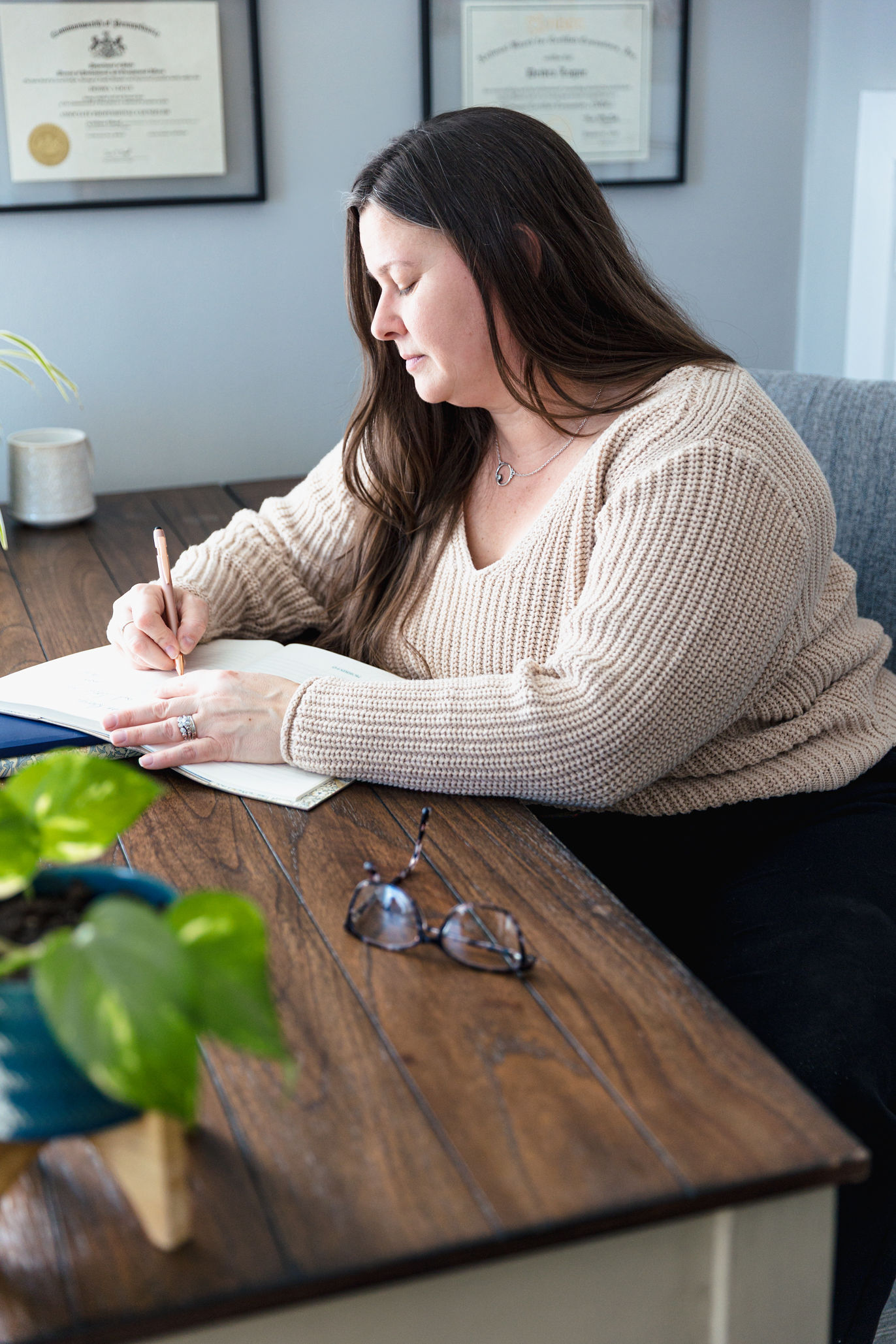 A woman with long brown hair sits at a wooden desk, writing in a notebook. She wears a beige sweater. Glasses and a small potted plant are on the desk, while diplomas related to family therapy hang on the wall in the background.