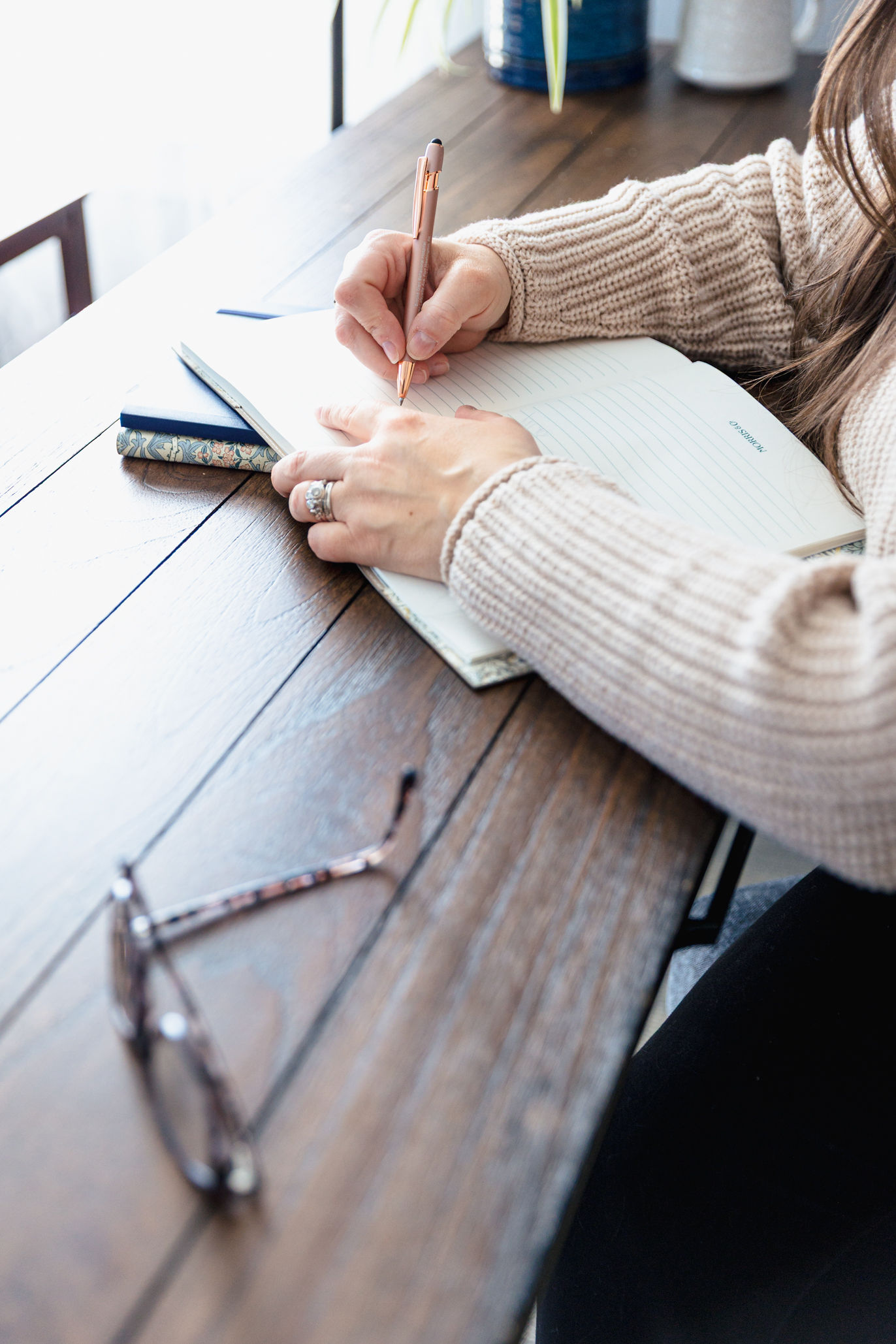 A person wearing a beige sweater writes in a lined notebook on a wooden table, possibly jotting down thoughts from couples therapy. A phone, eyeglasses, and pen are nearby as bright natural light streams in from the side.