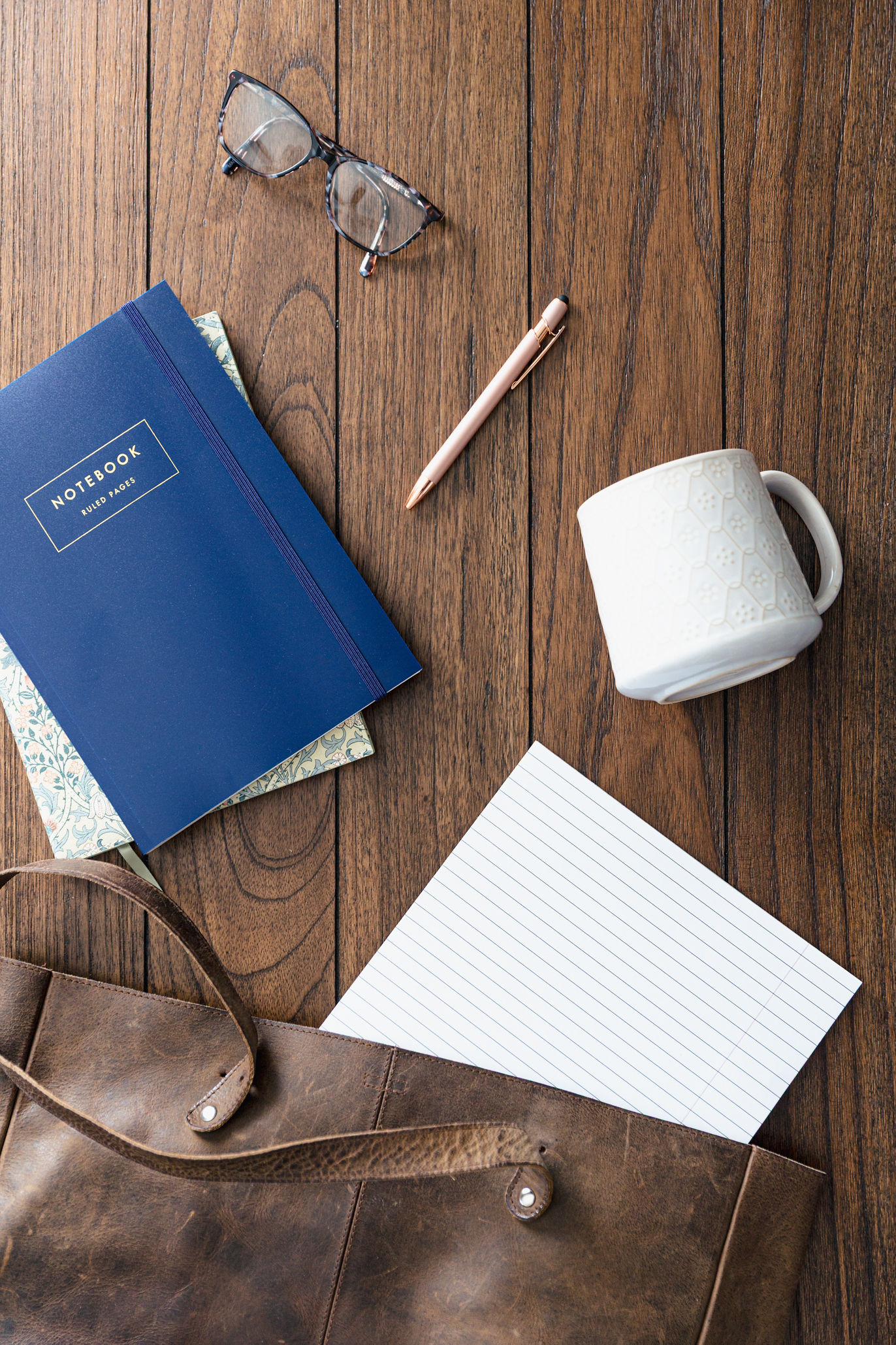 A pair of glasses, two notebooks, a pen, a white mug, a sheet of lined paper, and a brown leather bag are arranged on a wooden surface—tools often found in couples therapy sessions.