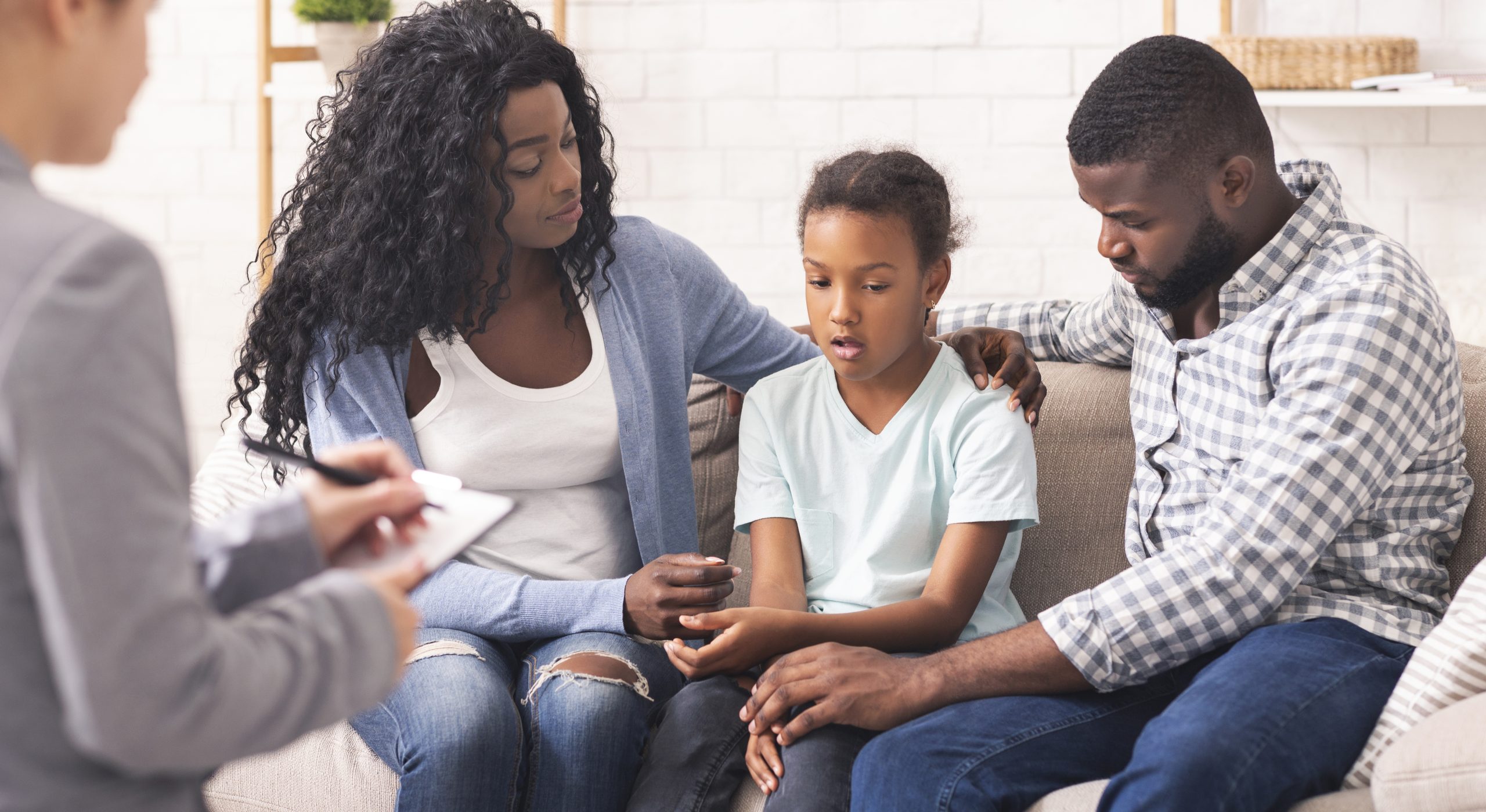 A young girl sits between two adults on a couch, looking down while they comfort her. Another person nearby, possibly a therapist, takes notes. The setting appears calm and supportive, suggesting a family therapy session.